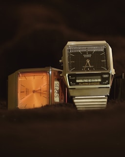 Two digital wristwatches are displayed with an emphasis on their vintage designs. The watch on the left has an orange face with a simple, classic design, while the watch on the right features a black face with a digital screen displaying the time, along with additional features. Both have metallic casings with a retro aesthetic.