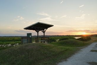 A wooden shelter is positioned on a grassy mound overlooking a scenic landscape. In the background, the sun is setting on the horizon, casting a warm glow over a river and surrounding greenery. A dirt path leads up to the shelter, adding to the natural and tranquil setting.