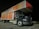 A large delivery truck is parked in a lot at night. The side of the truck features branding with the words 'Furnitureland South' and 'Let's Create Home' in bold, white lettering on a textured red and wood-patterned background. The front cab of the truck is black with silver accents and visible parking lines on the ground.