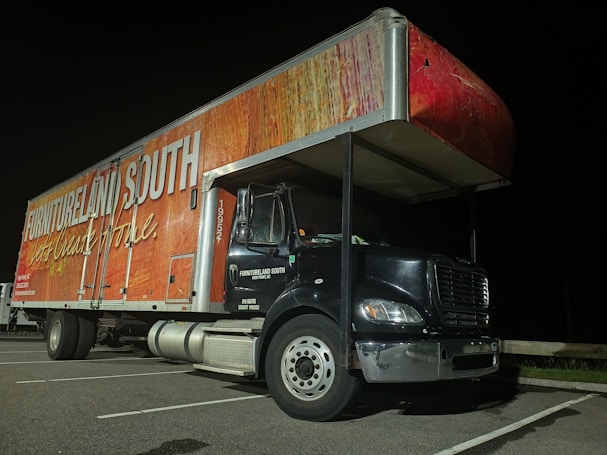 A large delivery truck is parked in a lot at night. The side of the truck features branding with the words 'Furnitureland South' and 'Let's Create Home' in bold, white lettering on a textured red and wood-patterned background. The front cab of the truck is black with silver accents and visible parking lines on the ground.