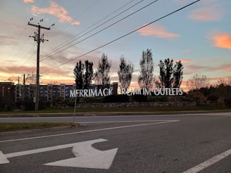 A large roadside sign for Merrimack Premium Outlets is prominently displayed against a backdrop of a colorful sunset. The sky features hues of pink and orange with scattered clouds. Tall trees frame the sign on either side, and power lines stretch across the sky. A road passes in front of the sign with a painted left-turn arrow visible.