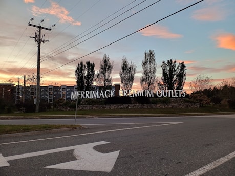 A large roadside sign for Merrimack Premium Outlets is prominently displayed against a backdrop of a colorful sunset. The sky features hues of pink and orange with scattered clouds. Tall trees frame the sign on either side, and power lines stretch across the sky. A road passes in front of the sign with a painted left-turn arrow visible.