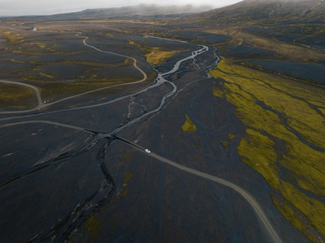 An aerial view of a vast landscape with meandering roads and a river cutting through a rocky terrain. The terrain is a mix of dark volcanic rock and patches of green vegetation. A small white vehicle is visible on one of the roads, accentuating the scale of the environment.