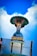 A statue stands on top of a globe, depicting a figure with outstretched hands under a stylized tree-like canopy. The structure is built above a white building and there are signs with text in an Indian script. The sky is clear and blue with a few clouds.