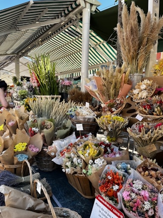 A lively outdoor market stall displays a variety of vibrant dried flowers in baskets and brown paper packaging. The arrangement includes colorful blooms such as yellow, red, pink, and white. Positioned under a green and white striped awning, different bunches are labeled with price tags.