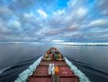 A cargo ship moves through a vast, calm ocean under a sky filled with dramatic clouds. The deck is loaded with colorful shipping containers, and the ship's wake creates a white trail in the blue water. The distant horizon features a low-lying landmass.