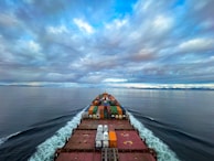 A cargo ship moves through a vast, calm ocean under a sky filled with dramatic clouds. The deck is loaded with colorful shipping containers, and the ship's wake creates a white trail in the blue water. The distant horizon features a low-lying landmass.