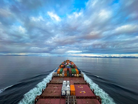 A cargo ship moves through a vast, calm ocean under a sky filled with dramatic clouds. The deck is loaded with colorful shipping containers, and the ship's wake creates a white trail in the blue water. The distant horizon features a low-lying landmass.