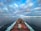 A cargo ship moves through a vast, calm ocean under a sky filled with dramatic clouds. The deck is loaded with colorful shipping containers, and the ship's wake creates a white trail in the blue water. The distant horizon features a low-lying landmass.