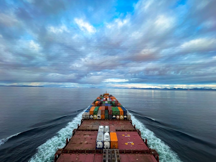 A cargo ship moves through a vast, calm ocean under a sky filled with dramatic clouds. The deck is loaded with colorful shipping containers, and the ship's wake creates a white trail in the blue water. The distant horizon features a low-lying landmass.