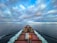A cargo ship moves through a vast, calm ocean under a sky filled with dramatic clouds. The deck is loaded with colorful shipping containers, and the ship's wake creates a white trail in the blue water. The distant horizon features a low-lying landmass.