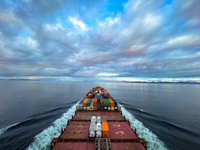 A cargo ship moves through a vast, calm ocean under a sky filled with dramatic clouds. The deck is loaded with colorful shipping containers, and the ship's wake creates a white trail in the blue water. The distant horizon features a low-lying landmass.