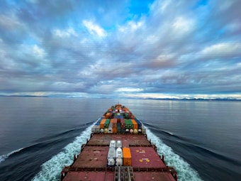 A cargo ship moves through a vast, calm ocean under a sky filled with dramatic clouds. The deck is loaded with colorful shipping containers, and the ship's wake creates a white trail in the blue water. The distant horizon features a low-lying landmass.