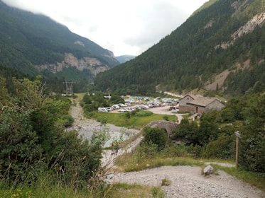 A picturesque valley surrounded by lush green mountains under a cloudy sky. A collection of parked cars is visible in a field near a rustic stone building. Dense forests cover the slopes of the mountains, with a rocky pathway leading through the vegetation.