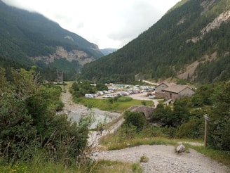 A picturesque valley surrounded by lush green mountains under a cloudy sky. A collection of parked cars is visible in a field near a rustic stone building. Dense forests cover the slopes of the mountains, with a rocky pathway leading through the vegetation.