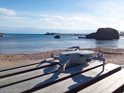 A drone is placed on a wooden surface at a beachside location. The clear blue sky and calm sea form the background, with boats visible near the horizon. Large rocks are also seen on the right side.