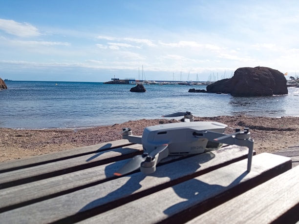 A drone is placed on a wooden surface at a beachside location. The clear blue sky and calm sea form the background, with boats visible near the horizon. Large rocks are also seen on the right side.