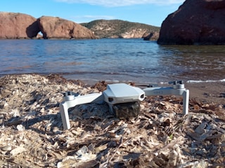 A small white drone is resting on a beach with dried seaweed and sand in the foreground. The beach is adjacent to a calm body of water, and in the background, there are rocky formations, with one prominent arch-like rock feature. The sky above is clear with minimal clouds.