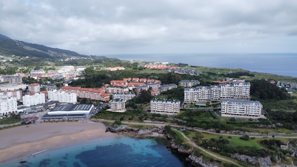 A coastal town with a variety of buildings clustered near the shoreline, surrounded by a lush green landscape. A sandy beach leads to clear, azure waters, while residential buildings vary from modern high-rises to traditional style houses. The sky is overcast, creating a muted ambiance.