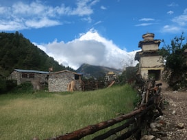 A rustic, stone-built house is surrounded by lush greenery and a wooden fence in a mountainous rural area. A prominent pagoda-style structure stands nearby, with a rugged, dirt path leading towards it. In the backdrop, a snow-capped mountain peak towers above the clouds, framed by a blue sky with scattered clouds.