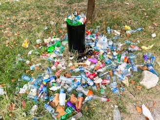 A black trash can is surrounded by a large quantity of litter, including plastic bottles, cans, and other waste items scattered on a grassy field. The scene conveys neglect and environmental pollution with fallen leaves adding to the untidiness.