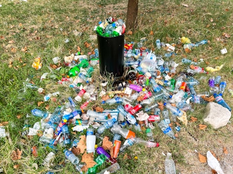 A black trash can is surrounded by a large quantity of litter, including plastic bottles, cans, and other waste items scattered on a grassy field. The scene conveys neglect and environmental pollution with fallen leaves adding to the untidiness.