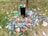 A black trash can is surrounded by a large quantity of litter, including plastic bottles, cans, and other waste items scattered on a grassy field. The scene conveys neglect and environmental pollution with fallen leaves adding to the untidiness.