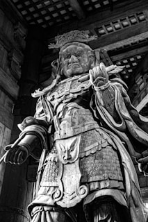A wooden sculpture of a fierce warrior adorned with intricate armor, including a detailed breastplate and a crown. The figure holds a staff and displays an intense expression, reflecting a sense of authority and power. The craftsmanship of the carving emphasizes the textures of the wood and the elaborate designs on the armor.