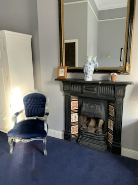 A room featuring an elegant black fireplace with decorative tiles, beneath a large gilded mirror. A blue and white vase sits atop the fireplace mantel, alongside a small pot and a picture frame. A plush, blue velvet chair with ornate silver detailing is positioned nearby. The room is decorated in soft grey tones with a dark blue carpet.