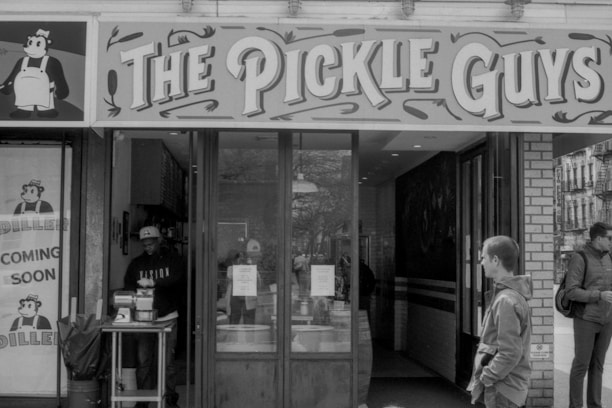 A storefront with a sign reading 'The Pickle Guys' is visible. An illustrated character in a chef outfit is part of the branding. A person is standing inside near a counter, and a few others are outside walking by. There's a 'Coming Soon' sign featuring the same illustrated character.