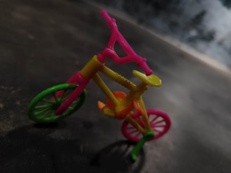 A miniature toy bicycle with vibrant colors, including pink, yellow, green, and orange. The bicycle is positioned at an angle on a dark surface, with a blurred background that gives a sense of depth.