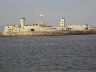 A concrete fort structure stands on a body of water, with angular, geometric lines and minimal windows. Two small towers are visible at both ends. A flagpole with a red and white flag is central on the fort. The water is calm, and the sky above is cloudy but bright.