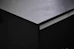 A close-up view of a dark, matte kitchen countertop. The surface texture appears smooth with subtle light reflections and markings. The countertop is part of a cabinet unit with clean lines and a modern design.