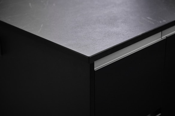 A close-up view of a dark, matte kitchen countertop. The surface texture appears smooth with subtle light reflections and markings. The countertop is part of a cabinet unit with clean lines and a modern design.