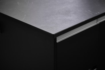 A close-up view of a dark, matte kitchen countertop. The surface texture appears smooth with subtle light reflections and markings. The countertop is part of a cabinet unit with clean lines and a modern design.