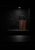 A set of knives is arranged in a wooden knife block, illuminated by a focused overhead light against a dark, marbled background. This minimalist setting highlights the polished surfaces of the knives, giving the scene a sleek and modern aesthetic.