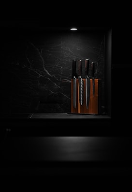A set of knives is arranged in a wooden knife block, illuminated by a focused overhead light against a dark, marbled background. This minimalist setting highlights the polished surfaces of the knives, giving the scene a sleek and modern aesthetic.