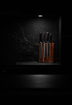 A set of knives is arranged in a wooden knife block, illuminated by a focused overhead light against a dark, marbled background. This minimalist setting highlights the polished surfaces of the knives, giving the scene a sleek and modern aesthetic.