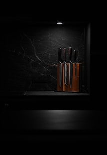 A set of knives is arranged in a wooden knife block, illuminated by a focused overhead light against a dark, marbled background. This minimalist setting highlights the polished surfaces of the knives, giving the scene a sleek and modern aesthetic.