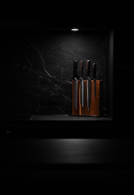 A set of knives is arranged in a wooden knife block, illuminated by a focused overhead light against a dark, marbled background. This minimalist setting highlights the polished surfaces of the knives, giving the scene a sleek and modern aesthetic.