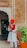 A man dressed in a traditional white and red outfit with a red turban stands holding a staff at the entrance of a building. The building has an arched doorway and a faded orange wall with white decorative accents. There is a potted plant with green foliage to his right.