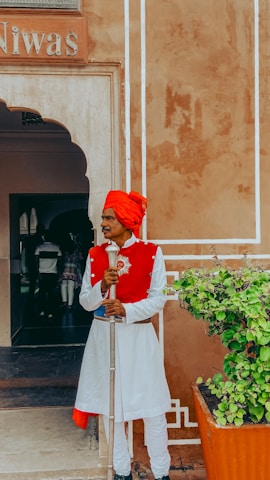 A man dressed in a traditional white and red outfit with a red turban stands holding a staff at the entrance of a building. The building has an arched doorway and a faded orange wall with white decorative accents. There is a potted plant with green foliage to his right.