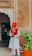 A man dressed in a traditional white and red outfit with a red turban stands holding a staff at the entrance of a building. The building has an arched doorway and a faded orange wall with white decorative accents. There is a potted plant with green foliage to his right.