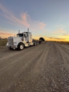 A large semi-truck with a trailer is parked on a gravel road. The trailer carries a cylindrical object. The sky is expansive with a backdrop of a colorful sunset, casting orange and pink hues against the wispy clouds.