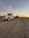 A large semi-truck with a trailer is parked on a gravel road. The trailer carries a cylindrical object. The sky is expansive with a backdrop of a colorful sunset, casting orange and pink hues against the wispy clouds.