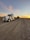 A large semi-truck with a trailer is parked on a gravel road. The trailer carries a cylindrical object. The sky is expansive with a backdrop of a colorful sunset, casting orange and pink hues against the wispy clouds.