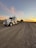 A large semi-truck with a trailer is parked on a gravel road. The trailer carries a cylindrical object. The sky is expansive with a backdrop of a colorful sunset, casting orange and pink hues against the wispy clouds.