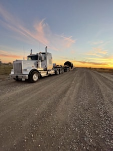 A large semi-truck with a trailer is parked on a gravel road. The trailer carries a cylindrical object. The sky is expansive with a backdrop of a colorful sunset, casting orange and pink hues against the wispy clouds.