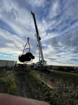 A large crane is lifting a hefty object, possibly industrial equipment, in an open field. The sky is partly cloudy, with patches of blue showing through. There's a dirt path surrounded by tall grass leading to the crane. A worker in a reflective vest stands near the crane, monitoring the operation. In the background, shipping containers and trailers are visible.