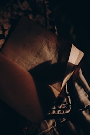 An open book with text on a page partially covered by shadows. The lighting is dim and creates a mysterious, intimate atmosphere, with fabrics underneath adding texture.